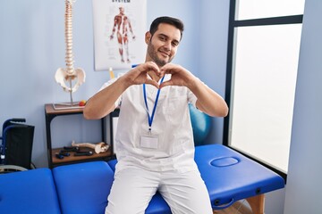 Sticker - Young hispanic man with beard working at pain recovery clinic smiling in love doing heart symbol shape with hands. romantic concept.