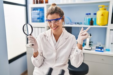 Sticker - Young woman working at scientist laboratory holding magnifying glass smiling happy pointing with hand and finger to the side