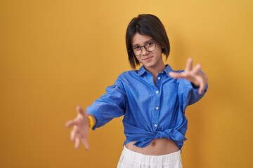 Poster - Young girl standing over yellow background looking at the camera smiling with open arms for hug. cheerful expression embracing happiness.