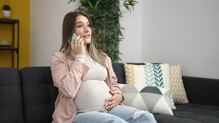 Canvas Print - Young pregnant woman talking on smartphone touching belly at home