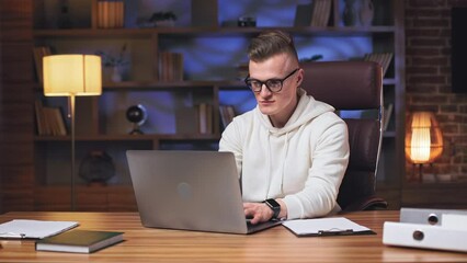 Casually dressed caucasian male programmer wearing glasses working closely on writing and testing new codes in spacious office. Focused man sitting in comfy rolling chair and using personal laptop.