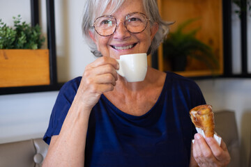 Wall Mural - Happy senior woman with eyeglasses sitting in cafeteria enjoying coffee cup espresso and a sweet cake