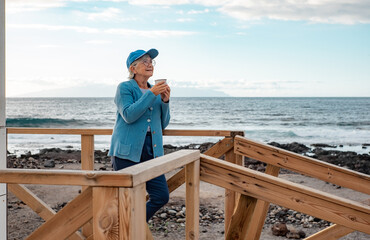 Smiling senior woman in blue standing outdoors at the beach in sunset light holding a coffee cup looking away. Happy pensioner woman enjoying free lifestyle in retirement