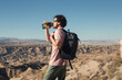Side shot of young man hiking in desert and hot area or landscape. Look at sun in sport sunglasses, drink water from see through water hydration flask, carry backpack with hiking gear