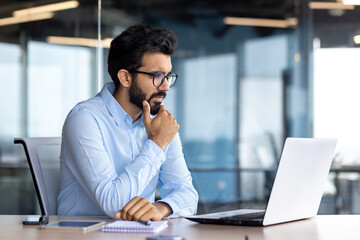 Serious thinking businessman inside office at workplace, mature Indian man in shirt working on laptop, programmer in glasses thinking about solving technical problem