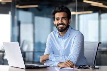 Portrait of happy and successful businessman, indian man smiling and looking at camera, satisfied with achievement results man working inside office building using laptop at work