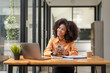 © Wasana - African American woman sitting in front of a laptop and holding a mobile phone, looking outside.