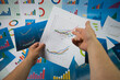 Ręka wskazująca wykres na białej kartce papieru trzymanej w dłoni na tle plansz z wykresami.