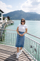 Wall Mural - Portrait of young and stylish woman stand on bridge and smile. Background of beautiful lake. Clear blue water.