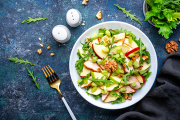 Waldorf salad with fresh apple, celery, lettuce, grilled chicken fillet, arugula and walnuts on plate, blue table background, top view