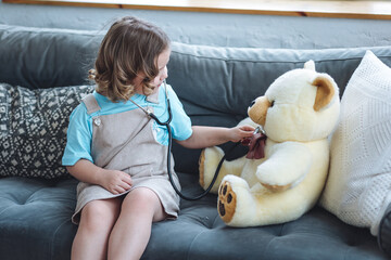 cute little girl in a beautiful dress paying doctor at home with her teddy bear, listening to heartb