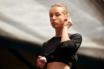 Wall Mural - A cute young athlete girl takes out wireless headphones from a case and puts them in the ears. Attractive blonde in a black t-shirt listens to music and prepares for a workout. Active lifestyle.