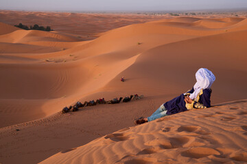 Berber man, an original inhabitant of North Africa, in traditional clothes in its natural habitat in Sahara Desert. Berber resting on an orange dune at the sunset. Camels caravan at the background.