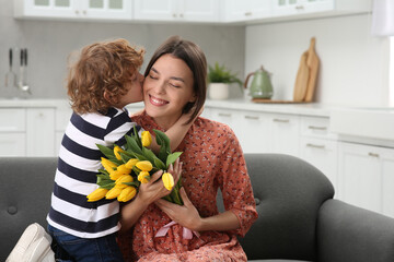 Sticker - Little son kissing and congratulating his mom with Mother`s day at home. Woman holding bouquet of yellow tulips