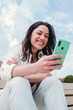 © Jose Calsina - Vertical portrait of young adult woman watching the social media on a cellphone app sitting outdoors. Carefree female smiling and having fun texting messages with a smartphone. Lady using mobile phone