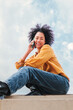 © Jose Calsina - Vertical portrait of young adult hispanic woman with curly hair having a cellphone call conversation sitting under blue sky with clouds. Happy latin girl talking by phone, at background the skyline