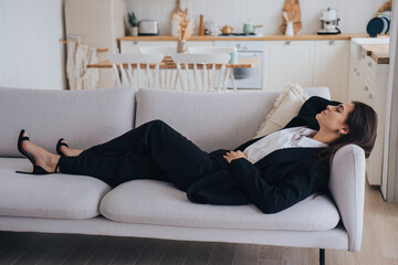 Tired brunette businesswoman in black suit laying on couch eyes closed at home relaxing after hard working. Lack of energy, exhausted employee needs rest. Beautiful girl sleeping on sofa at home.