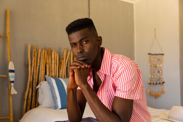 Portrait of thoughtful biracial man sitting on bed at summer beach house