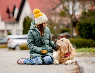 Wall Mural - Preteen child girl sitting with golden retriever dog on asphalt at autumn street wearing hat and warm jacket. Pretty kid petting purebred pet doggy labrador outdoors at city