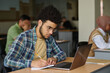 © AnnaStills - Young man sitting at desk with laptop and making notes in notepad, he learning language in class