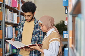 Couple of multiethnic students standing near the bookcase and reading book together in library