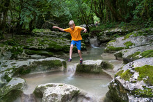 Active Boy Jumps From Rock To Rock On A Calm Mountain River Hidden In A Dense Forest