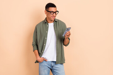 Photo of good mood clever guy dressed khaki shirt glasses looking at smartphone hold arm in pocket isolated on pastel color background