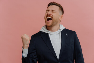 Triumphant unshaven man celebrates success, raising clenched fist, screaming in joy against pink background