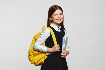 Wall Mural - Carefree pupil girl with yellow backpack smiling and embracing notebooks during studies against white background. Back to school concept