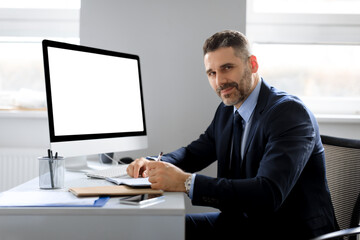 Poster - Cheerful middle aged businessman sitting at workplace with computer with blank screen, free space, mockup template