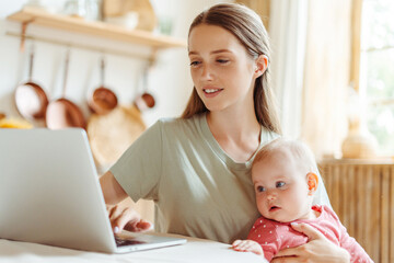 Young smiling woman, freelancer holding cute baby using laptop working online from home, watching videos. Parenthood concept 