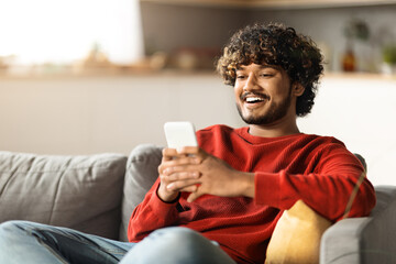 Sticker - Cheerful Indian Man Messaging On Smartphone While Relaxing On Couch At Home