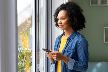 Happy biracial woman using smartphone and looking out window at home