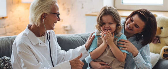 Kind senior female paediatrician doctor examining little child sitting on mother's laps, during home visit or clinic check up. Concept of healthcare, medical assistance, insurance. Banner