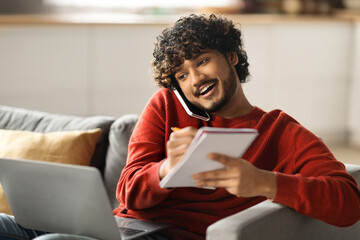 Wall Mural - Work From Home. Young Indian Man Talking On Cellphone And Taking Notes