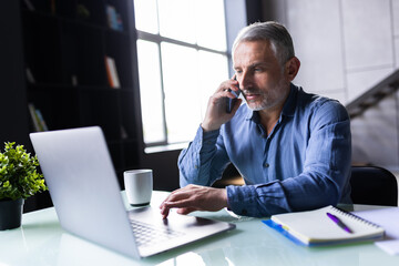 Mid adult business professional sitting at table with laptop and using mobile phone.