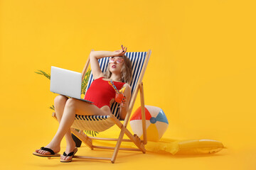 Wall Mural - Upset young woman with cocktail and laptop in deck chair on yellow background