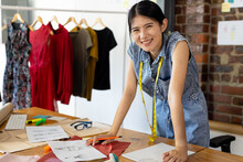 Portrait Of Asian Female Fashion Designer Wearing Tape Measure Leaning On Desk Smiling At Studio