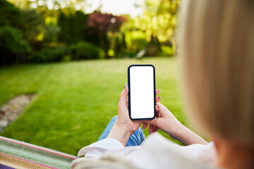 close up of woman using phone in garden sitting in hammock with blank mock up screen