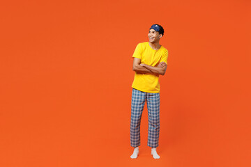 Wall Mural - Full body smiling cheerful fun happy young man wear pyjamas jam sleep eye mask rest relax at home look aside on area isolated on plain orange background studio portrait. Good mood night nap concept.