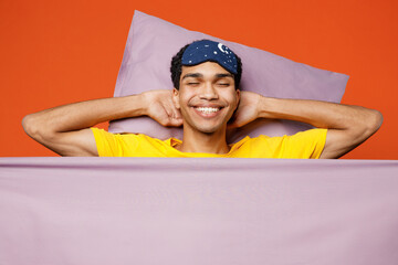 Wall Mural - Calm young fun man wearing pyjamas jam sleep eye mask rest relax at home lies wrap covered under blanket duvet close eyes isolated on plain orange color background studio. Good mood night nap concept.