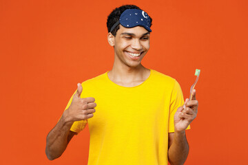 Wall Mural - Calm smiling young man wear pyjamas jam sleep eye mask rest relax at home brushing teeth show thumb up isolated on plain orange background studio portrait. Good mood night nap, daily routine concept.