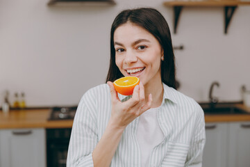 Young smiling happy housewife latin woman wear casual clothes striped shirt eating biting half of orange fruit look camera sit at table in light kitchen at home alone. Lifestyle cooking food concept.