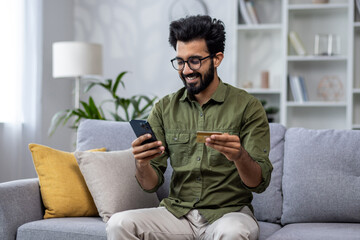 Happy and satisfied man shopping online sitting on sofa at home, hispanic customer holding bank credit card and phone, using online shopping app.