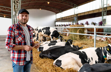 Portrait young adult farmer holding digital tablet in hands and smiling against background of cows in paddock in livestock farm.