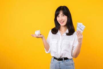 Poster - Portrait beautiful young business asian woman wearing white shirt and denim jean with a lot of cash money and piggy bank isolated on yellow background. Wealth money saving, finance investment concept.