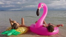Two Girlfriends Lie On The Beach On Flamingo And Pineapple-shaped Inflatable Laps And Talk To Each Other. Having A Nice Time On The Beach During A Weekend Vacation.