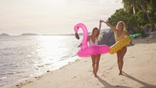 Two Girlfriends Walk Along The Beach Dancing In The Sunset Time Holding Hands After Swimming In The Sea With Pineapple And Flamingo Shaped Inflatable Circles. The Women Enjoy Their Vacation