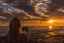 Woman Taking Photos Of The Sunset On The Beach With Mobile Phone. Summer, Travel And Technology Concept.