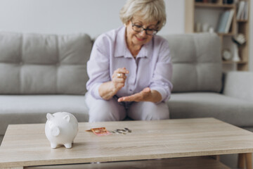 savings, money, annuity insurance, retirement and people concept - smiling senior woman putting coins into piggy bank at home